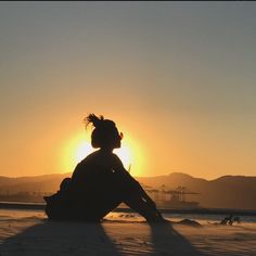 a woman is sitting on the beach watching the sun go down in the distance with her hair blowing in the wind