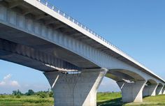 the underside of a bridge over a body of water with grass and trees in the background