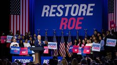 a man standing at a podium in front of a crowd with signs reading restore roe