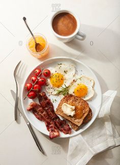 breakfast plate with eggs, bacon, toast and tomato on white tablecloth next to cup of coffee