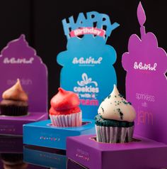 three cupcakes with frosting and sprinkles are displayed in purple boxes