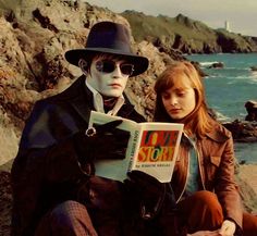 two people are sitting on the rocks by the ocean and reading a book, one is wearing a black hat