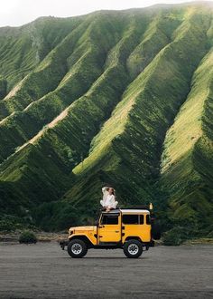 two people sitting on top of a yellow jeep in front of a green mountain range