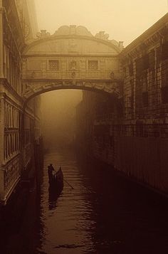 two people in a boat on a canal under an overpass with buildings around it