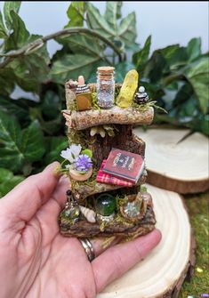 a hand holding a miniature fairy house made out of tree stumps and rocks with books on it