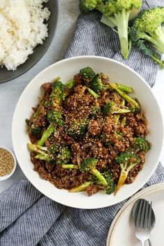 a white bowl filled with meat and broccoli next to rice on a table