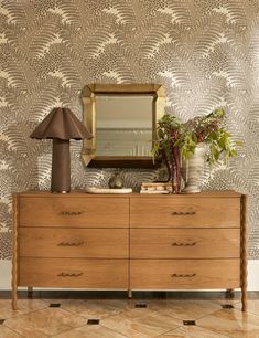 a dresser with a mirror, lamp and plants on it in front of a wallpapered background