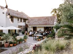 people are sitting at tables outside in front of a white building with lots of greenery