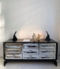 a black and silver dresser with two sculptures on it's top, in front of a white wall