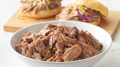 a white bowl filled with pulled pork next to a bun on a cutting board and another sandwich in the background