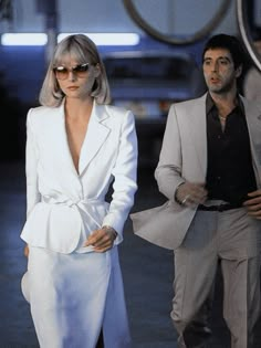 a woman in a white suit and sunglasses walking next to a man with grey hair