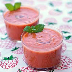 two glasses filled with red liquid sitting on top of a white table covered in green leaves