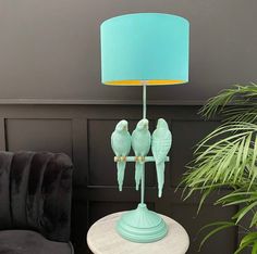 two green birds sitting on a table with a blue lamp in front of it and a potted plant next to it