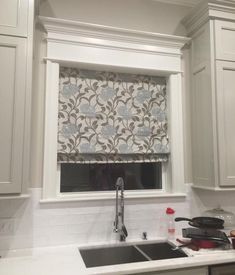 a kitchen with white cabinets and a window covered in roman blind shades over the sink