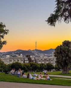 people are sitting on the grass in front of some buildings and trees, while the sun is setting