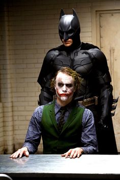 two men dressed as batman and the dark knight sit at a table in front of a brick wall