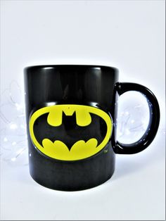 a black and yellow batman mug sitting on top of a white table next to a string of lights