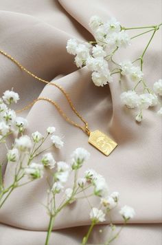 a gold necklace with a medallion on it sitting next to white flowers and a cloth