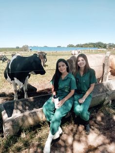 two women in scrubs are sitting on a fence with cows