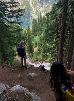 two people with backpacks are standing on a trail in the woods looking at mountains