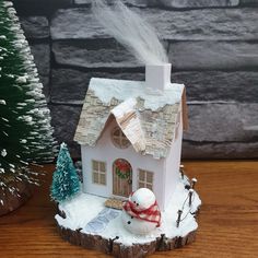 a small white house with snow on the roof and trees around it, next to a fake christmas tree