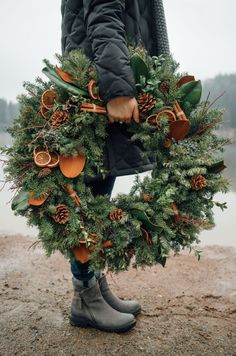 a person holding a wreath with pine cones and oranges on it in front of a body of water