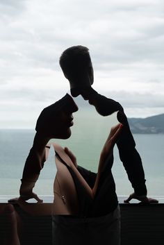 two people are standing in front of a window with the ocean and mountains behind them