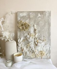 a white vase with flowers and candles on a bed next to a wall mounted painting