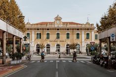 two people crossing the street in front of a large yellow building with green shutters