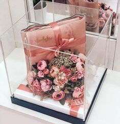 two clear boxes with pink flowers in them on top of a white table and one is holding a purse