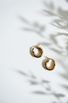 a pair of gold hoop earrings sitting on top of a white surface with shadows from the leaves