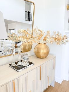 a gold vase with white flowers on top of a wooden table next to a mirror