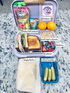 an open lunch box filled with food on top of a marble countertop next to oranges and cucumbers