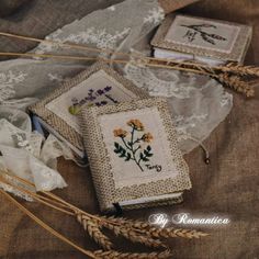 three cross stitch books with flowers on them sitting on some lace and burlap