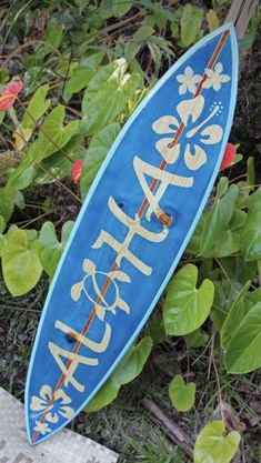 a blue surfboard sitting on the ground next to some green plants and flowers in front of it