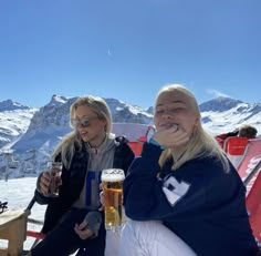 two women sitting on top of a snow covered ski slope drinking beer and smiling at the camera