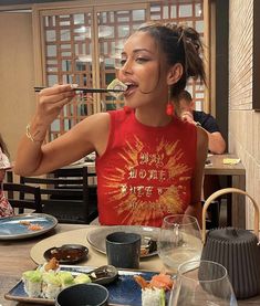 a woman sitting at a table eating food with chopsticks in her mouth