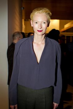 a woman with blonde hair standing in front of a wall wearing a purple blouse and black pants