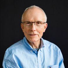 an older man with glasses and a blue shirt is looking at the camera while standing in front of a black background