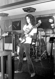 black and white photograph of a man playing guitar in a room with other instruments on the wall