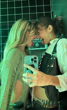 two women standing next to each other in front of a green tiled wall and one holding a cell phone