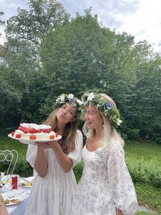 two women in white dresses holding a cake with strawberries on it and flowers around her head