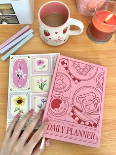 a person's hand on top of a pink planner next to a cup of coffee