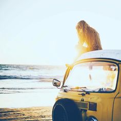 a woman sitting on the hood of a small yellow car at the ocean's edge