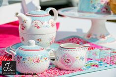 three tea pots and two cups are sitting on a colorful tablecloth with plates in the background