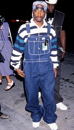 a man wearing overalls and a hat standing in front of other people at night