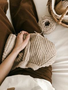 a person is knitting on a bed with brown and white blankets, two balls of twine are next to them