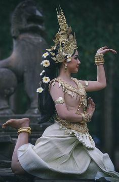 a woman dressed in white and gold sitting on the ground with her hands up to her chest