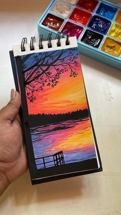 a hand holding a notebook with a sunset painting on it