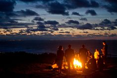 three people sitting around a campfire on the beach at night with fireworks in the sky